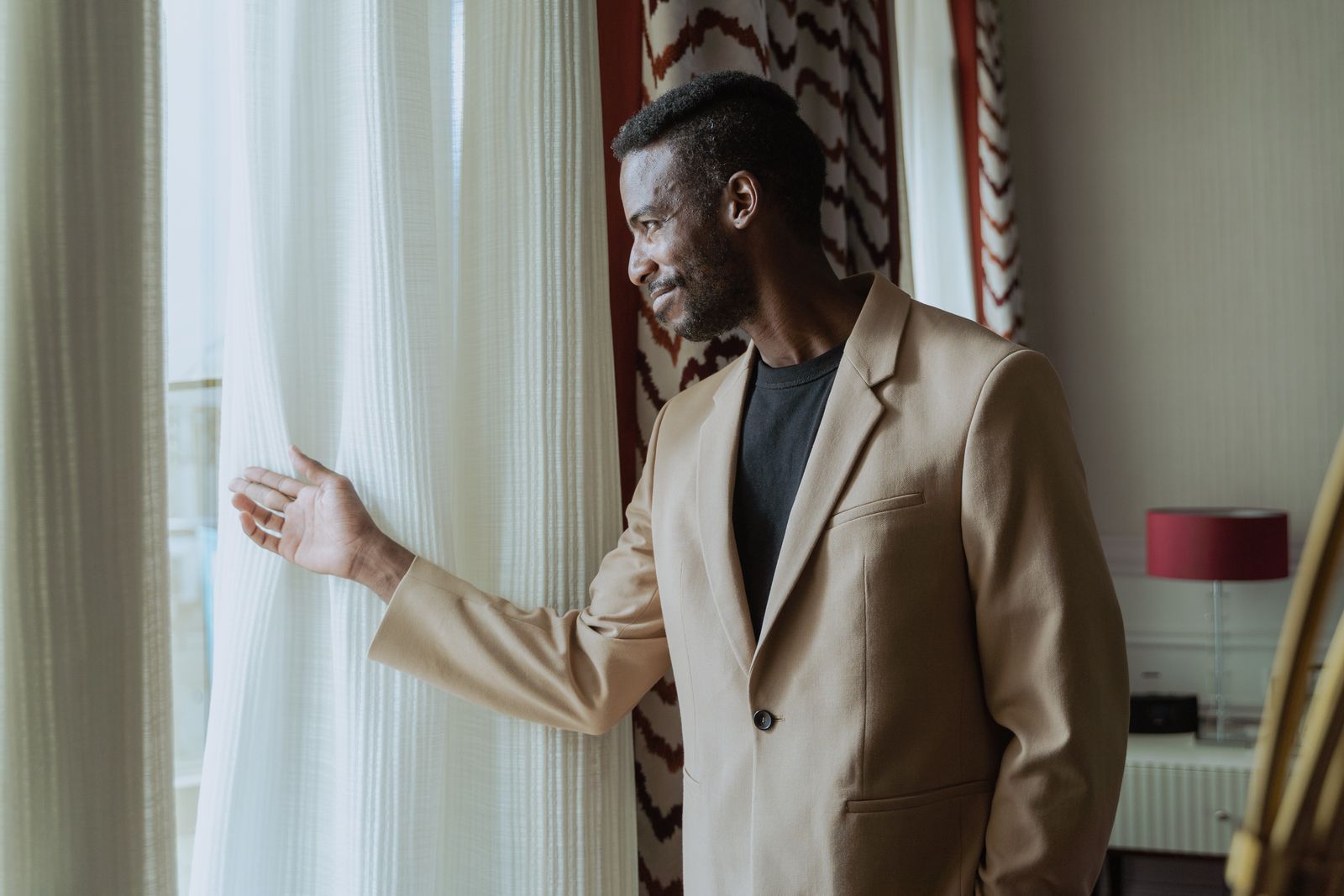 Un homme souriant, en costume beige, près de la fenêtre de son salon